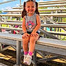 child, girl, smiling, pigtails, bleachers, outdoor, sunny, blue_sky, grass, sneakers, pink_socks, casual_clothing, sports, happy, summer, daytime, portrait, seated, metal_bench, playful
