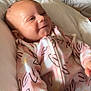 apparel, baby, child, cozy, cute, face, floral_pattern, happy, head, indoors, infant, lying_down, newborn, onesie, pink, portrait, skin, smile, soft_surface, young_child