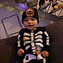 toddler, child, skeleton_costume, halloween, beanie, pumpkin, smile, couch, indoor, plush_toys, black_clothing, baby, cute, holiday, spooky, decorations, happy, person, young_child, festive