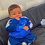 baby, child, blue_tracksuit, puma, socks, indoor, couch, leather, sitting, hand_in_mouth, cute, portrait, young_child, comfortable, clothing, person, face, expression, relaxed, home