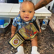 Asión is registered to the contest to win money with this photo: baby, casual, child, curious, door, floor, high_chair, home, indoor, kitchen, marble_countertop, person, plaid_shirt, plastic_container, smiling, table, tattooed_arm, water_bottle, white_socks, wwe_championship_belt