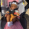 baby, child, stroller, headband, white_bow, floral_shirt, mustard_overalls, pink_cushion, safety_strap, sunlight, outdoor, infant, clothing, footwear, sock, person, seat, warning_label, relaxed, portrait