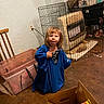 Oaklyn is registered to the contest to win money with this photo: toddler, child, blue_shirt, oversized_clothing, indoor, heater, wire_cage, plastic_bin, cardboard_box, floor, wall, electrical_outlet, curious_expression, standing, messy_room, toys, clutter, person, young_child, casual_clothing