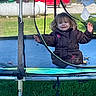 Oaklyn is registered to the contest to win money with this photo: child, trampoline, mesh_net, brown_coat, grass, outdoor, playground, toy, vehicle, truck, young_child, person, smiling, jacket, daylight, play, netting, parked_car, casual, fun