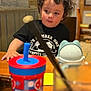Shivan joined the competition — help win amazing prizes! child, toddler, curly_hair, black_tshirt, soccer_ball, table, wooden_table, sippy_cup, toy, indoor, curious, face, person, cup, plastic, colorful, furniture, young_child, casual, seated