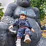 Shivan is registered to the contest to win money with this photo: toddler, child, statue, gorilla, outdoor, park, smiling, blue_outfit, cap, footwear, trees, greenery, playground, sculpture, person, happy, sitting, daylight, casual_clothing, nature