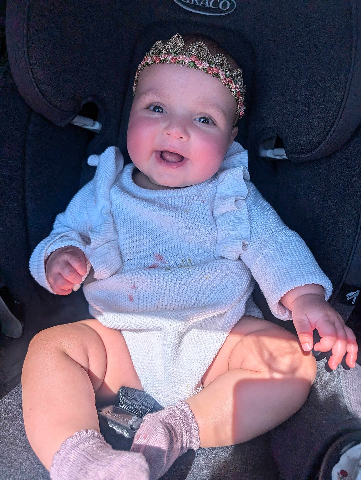 Dahlia is registered to the contest to win money with this photo: baby, child, car_seat, smiling, headband, knitted_clothing, pink_socks, rosy_cheeks, happy, infant, cute, sitting, sunlight, shadow, indoors, person, young_child, portrait, clothing, closeup