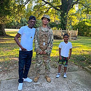 Jermaine is registered to the contest to win money with this photo: child, boy, adult, soldier, military_uniform, family, outdoor, park, backyard, tree, grass, deck, sunlight, portrait, standing, smiling, camouflage, cap, casual_clothing, group_photo