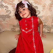 Aakifa is registered to the contest to win money with this photo: child, girl, red_dress, smile, ribbons, hair_accessory, couch, indoor, patterned_wallpaper, cheerful, portrait, cute, playful, home_interior, seated, young_child, decor, furniture, curtain, happy