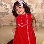 child, girl, red_dress, smile, ribbons, hair_accessory, couch, indoor, patterned_wallpaper, cheerful, portrait, cute, playful, home_interior, seated, young_child, decor, furniture, curtain, happy
