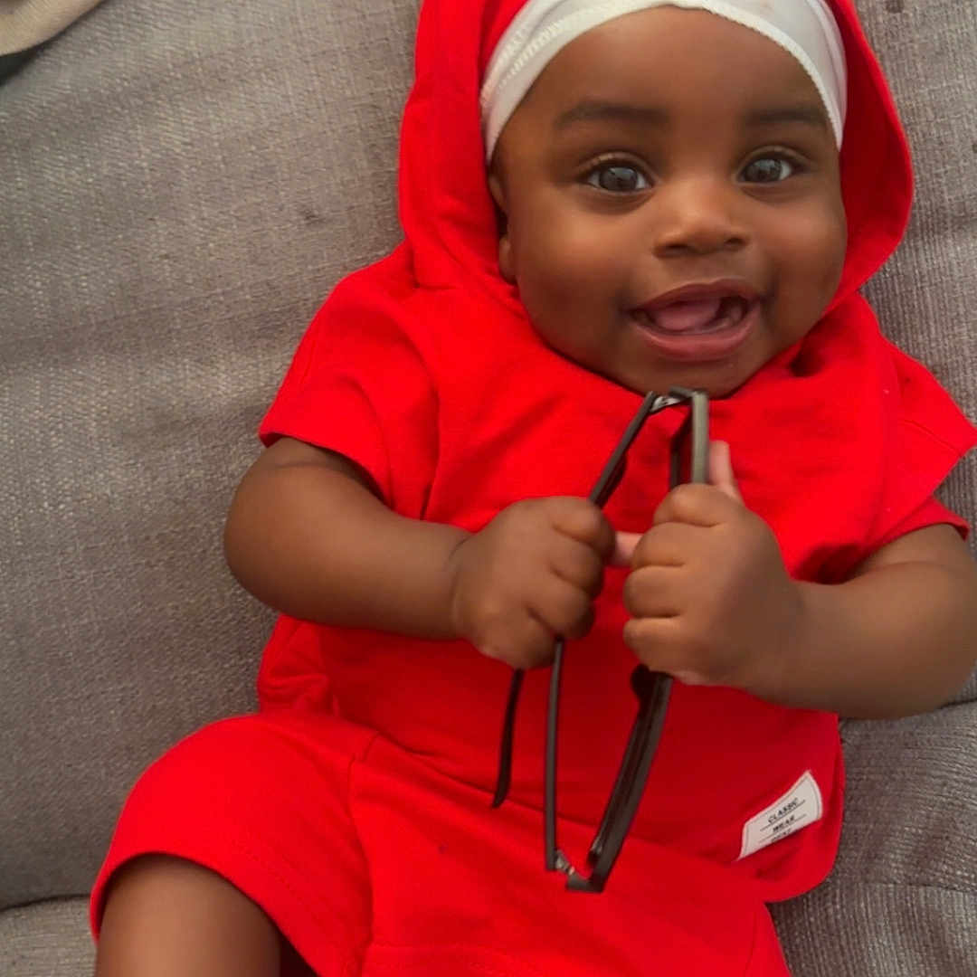 Zaccai is registered to the contest to win money with this photo: baby, casual, child, couch, cute, face, foot, hand, happy, headwear, holding_object, indoor, infant, one_person, portrait, red_clothing, relaxed, sitting, skin, smiling