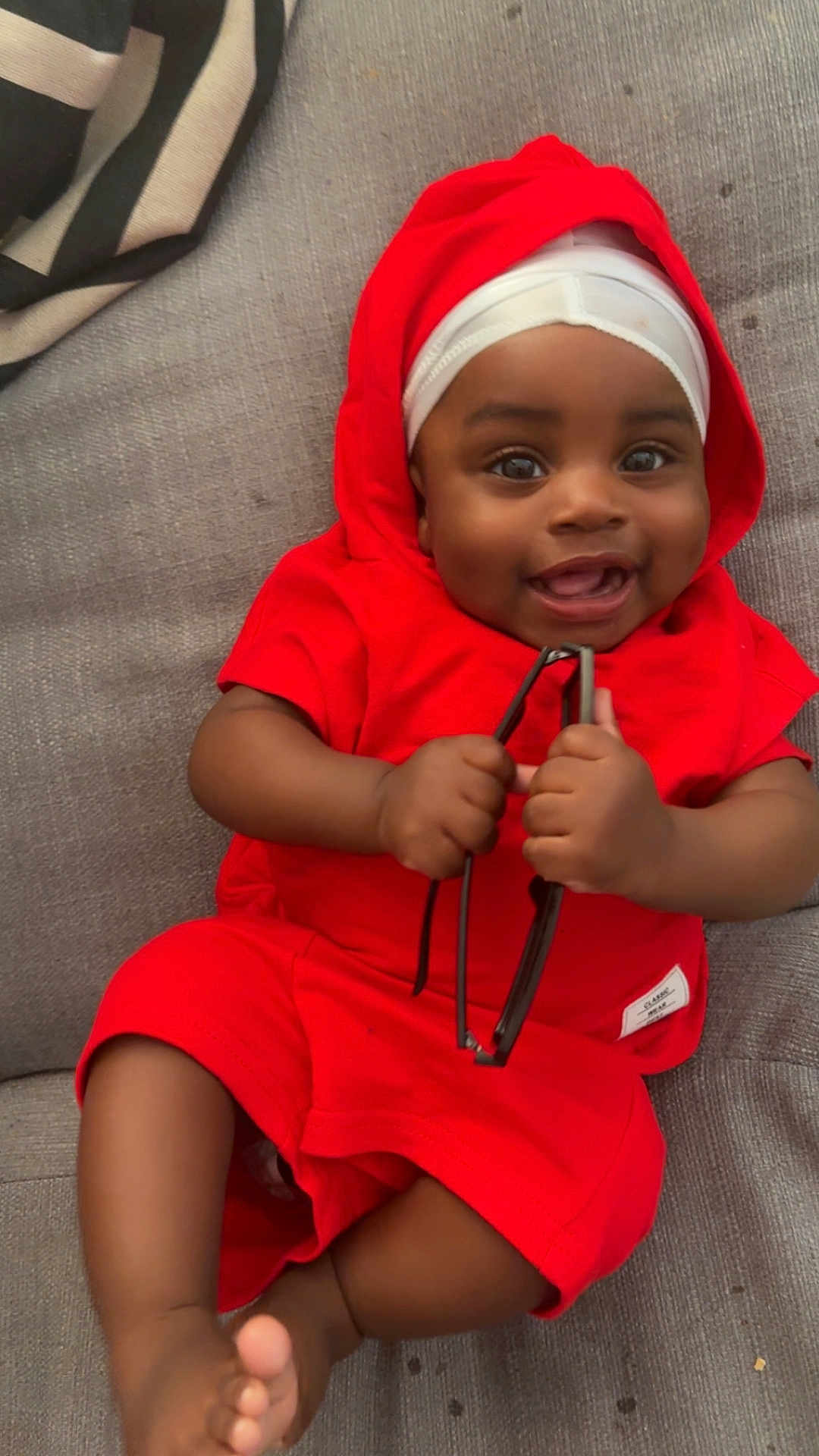 Zaccai is registered to the contest to win money with this photo: baby, child, smiling, red_clothing, couch, indoor, holding_object, happy, face, infant, foot, hand, headwear, portrait, one_person, casual, relaxed, cute, skin, sitting