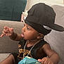 Zaccai is registered to the contest to win money with this photo: toddler, child, cap, black_clothing, chain_necklace, gray_couch, indoor, baby_wipes, toy, hand, foot, skin, face, sitting, curious, expression, living_room, furniture, person, casual
