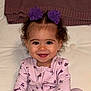 toddler, child, smiling, purple_flower, hair_clip, curly_hair, pink_onesie, sitting, indoor, blanket, happy, face, portrait, baby, cute, infant, apparel, person, cozy, background