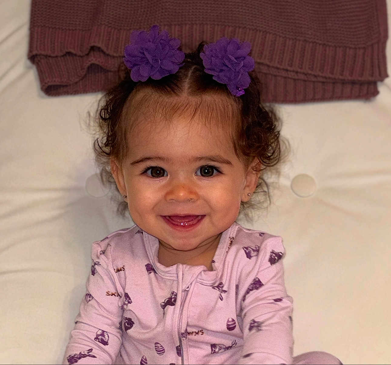 Legaci is registered to the contest to win money with this photo: toddler, child, smiling, purple_flower, hair_clip, curly_hair, pink_onesie, sitting, indoor, blanket, happy, face, portrait, baby, cute, infant, apparel, person, cozy, background