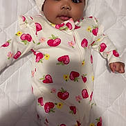 Malaysia is registered to the contest to win money with this photo: baby, infant, child, onesie, headband, strawberry_pattern, quilted_bed, lying_down, cute, adorable, sleepwear, curly_hair, dark_skin, portrait, indoors, soft_lighting, fist, looking_at_camera, white_background, comfortable