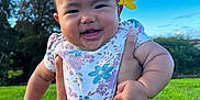 Hailey is registered to the contest to win money with this photo: baby, child, flower, smile, happy, outdoor, grass, blue_sky, onesie, arms, tattoo, person, nature, sunlight, portrait, cute, infant, holding, greenery, daytime