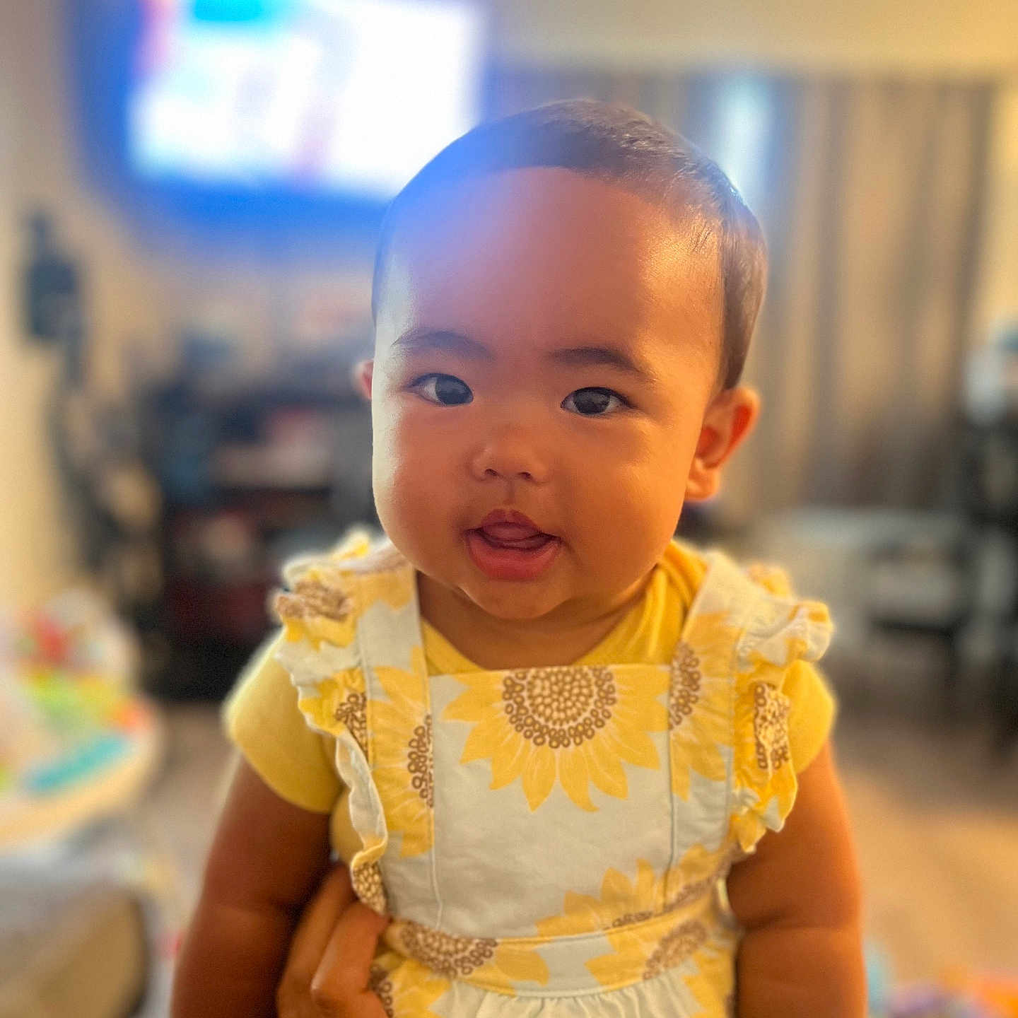 Hailey is registered to the contest to win money with this photo: baby, blurred_background, child, curtains, cute, face, hand, happy, holding, indoor, infant, living_room, person, portrait, round_face, smile, soft_light, sunflower_pattern, yellow_clothing, young_child