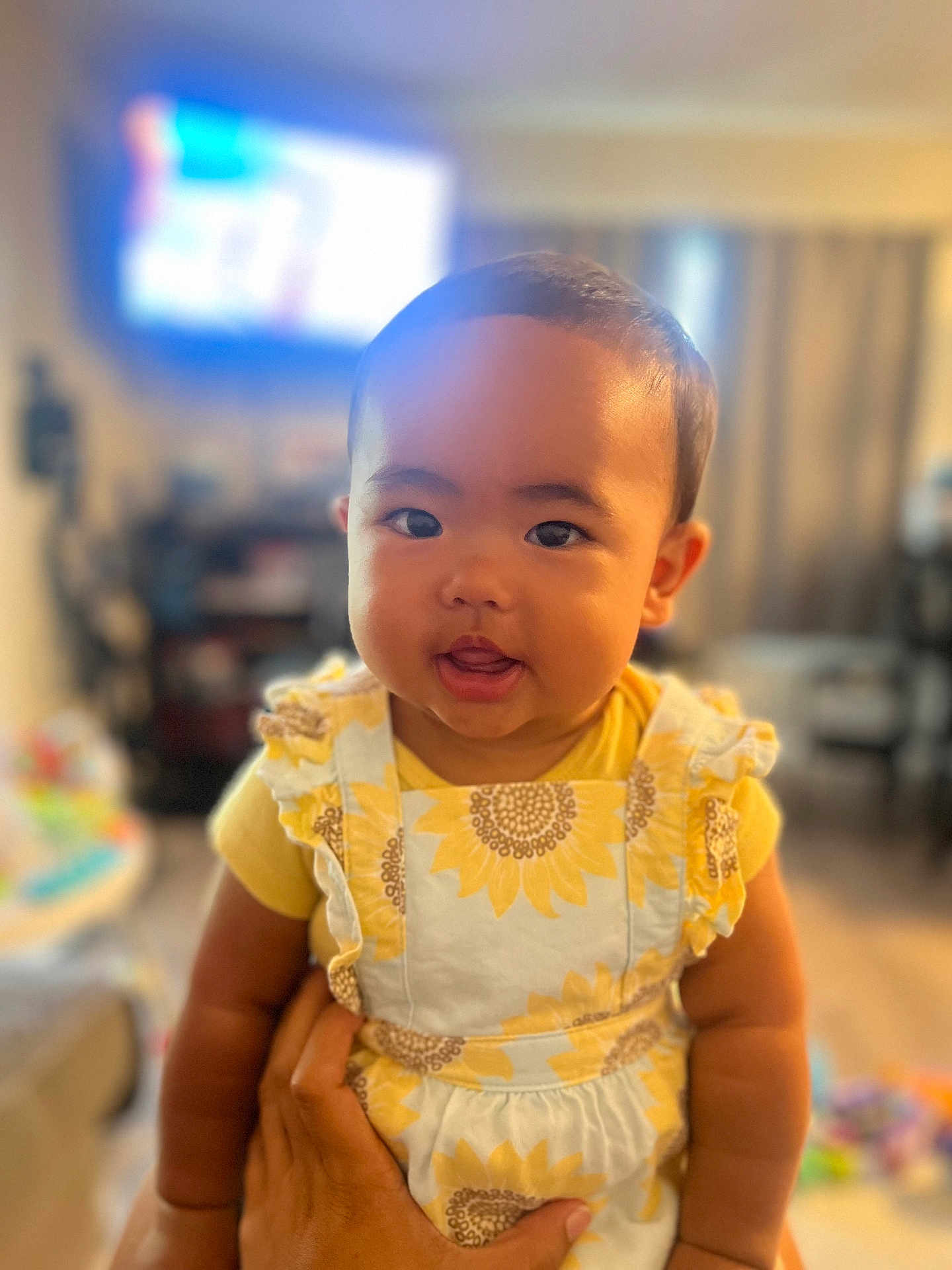 Hailey is registered to the contest to win money with this photo: baby, child, infant, face, smile, yellow_clothing, sunflower_pattern, hand, portrait, indoor, living_room, curtains, blurred_background, cute, young_child, holding, person, round_face, soft_light, happy