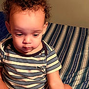 Ashley is registered to the contest to win money with this photo: baby, bed, bedroom, bedspread, child, cozy, curly_hair, eyes, face, indoors, looking_down, photography, portrait, puma_logo, sitting, striped_clothing, striped_pattern, t_shirt, toddler, warm_lighting