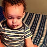 baby, bed, bedroom, bedspread, child, cozy, curly_hair, eyes, face, indoors, looking_down, photography, portrait, puma_logo, sitting, striped_clothing, striped_pattern, t_shirt, toddler, warm_lighting