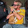 Chase is registered to the contest to win money with this photo: man, cat, orange_tabby, glasses, beard, yellow_shirt, shorts, chair, garage, vehicle, american_flag, tattoo, watch, pet, affection, indoor, casual, seated, human, animal