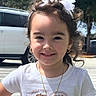 child, girl, smile, white_tshirt, heart_design, gold_necklace, gold_hoop_earrings, white_bow, hair_accessory, outdoor, sunny_day, trees, vehicle, sidewalk, person, portrait, casual_clothing, happy, cute, curly_hair