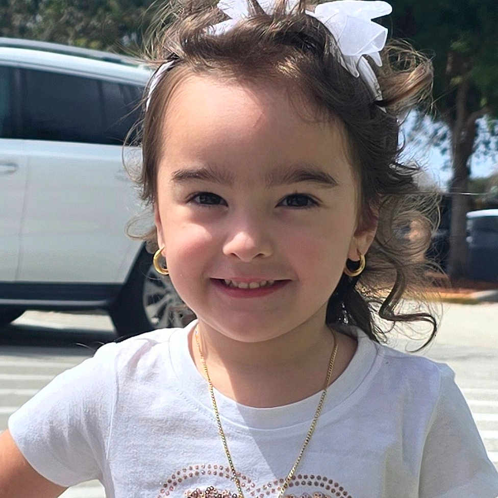 Brianna is registered to the contest to win money with this photo: child, girl, smile, white_tshirt, heart_design, gold_necklace, gold_hoop_earrings, white_bow, hair_accessory, outdoor, sunny_day, trees, vehicle, sidewalk, person, portrait, casual_clothing, happy, cute, curly_hair