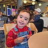Brianna is registered to the contest to win money with this photo: child, girl, pigtails, red_sweater, denim_overalls, smile, indoors, cafe, table, chair, people, woman, man, light, happy, portrait, casual_clothing, holding_object, seated, background