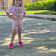 Brianna is registered to the contest to win money with this photo: child, girl, outdoor, sidewalk, smile, pink_clothing, bow, sneakers, shorts, tshirt, sunlight, greenery, house, residential, confident, posing, daytime, happy, casual, cute