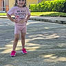 child, girl, outdoor, sidewalk, smile, pink_clothing, bow, sneakers, shorts, tshirt, sunlight, greenery, house, residential, confident, posing, daytime, happy, casual, cute