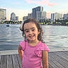 child, girl, smiling, pink_clothing, curly_hair, water, dock, cityscape, buildings, evening, outdoor, pose, happy, playful, urban, sky, light, nature, portrait, casual