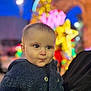 baby, child, face, blue_jacket, buttoned_clothing, curious_expression, holiday_lights, blurred_background, night, outdoor, person, portrait, cute, warm_clothing, festive, colorful_lights, winter, young_child, closeup, smile