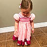 Paisley is registered to the contest to win money with this photo: child, girl, pink_dress, bow, hair_accessory, tile_floor, indoor, standing, looking_down, casual, young_child, flooring, wall, puffed_sleeves, collar, footwear, portrait, person, cute, shy
