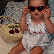 Paisley is registered to the contest to win money with this photo: toddler, child, sunglasses, red_sunglasses, white_outfit, cherry_pattern, bag, straw_bag, bed, pillow, indoor, cute, fashion, sitting, barefoot, person, portrait, young_child, hair_tied, playful