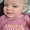 baby, infant, child, face, blonde_hair, pink_shirt, text_on_clothing, cousin_crew, blanket, soft_texture, closeup, portrait, cute, young_child, indoor, fabric, smile, head, skin, eyes