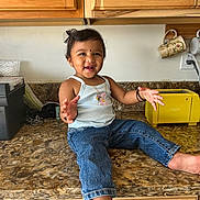 Shyna is registered to the contest to win money with this photo: appliance, barefoot, black_hair, child, countertop, face, hair, happy, home, indoor, jeans, jewelry, kitchen, mugs, person, smiling, tank_top, toaster, toddler, wooden_cabinets