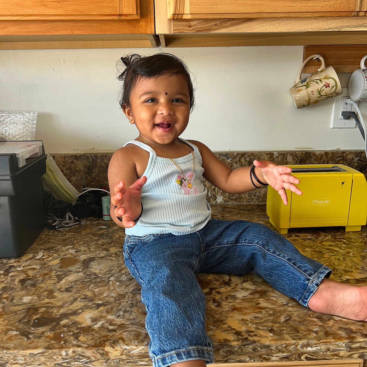 Shyna is registered to the contest to win money with this photo: appliance, barefoot, black_hair, child, countertop, face, hair, happy, home, indoor, jeans, jewelry, kitchen, mugs, person, smiling, tank_top, toaster, toddler, wooden_cabinets