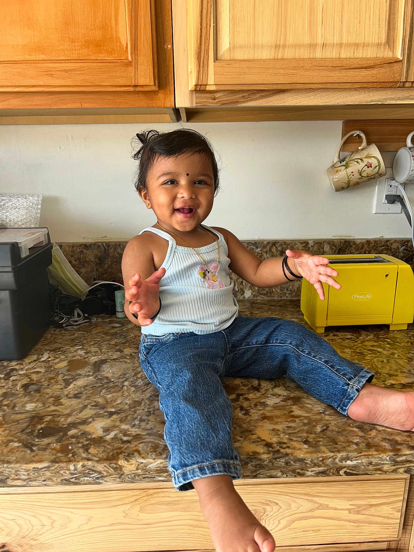 Shyna is registered to the contest to win money with this photo: appliance, barefoot, black_hair, child, countertop, face, hair, happy, home, indoor, jeans, jewelry, kitchen, mugs, person, smiling, tank_top, toaster, toddler, wooden_cabinets