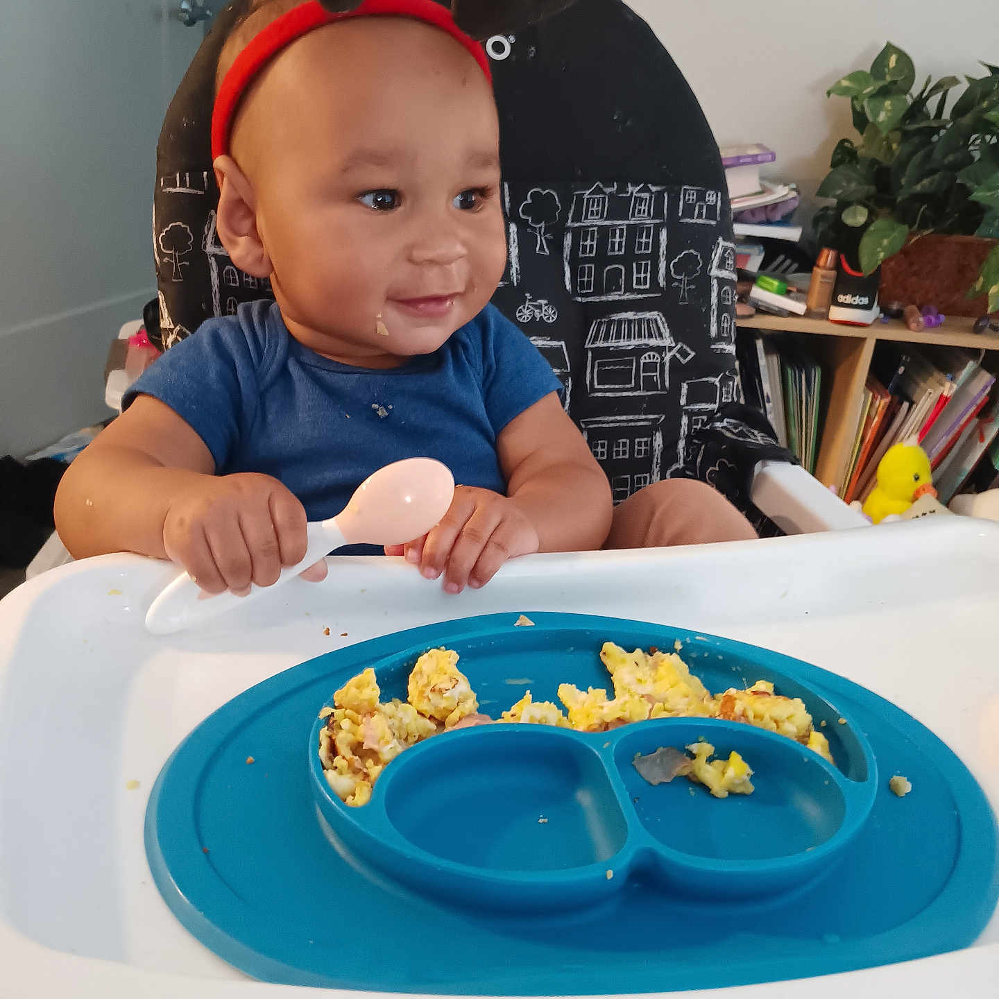Truce joined the competition — help win amazing prizes! baby, high_chair, spoon, food, scrambled_eggs, plate, reindeer_antlers, child, smiling, indoor, furniture, plant, books, toy_duck, blue_shirt, feeding_time, happy, face, person, home