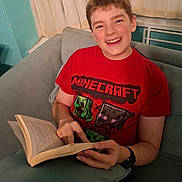 James is registered to the contest to win money with this photo: child, boy, smiling, portrait, indoor, sitting, couch, book, reading, minecraft, red_shirt, short_hair, happy, face, person, home, curtains, radiator, casual_clothing, photography
