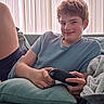 teenager, couch, controller, gaming, relaxed, smile, indoor, blanket, window, vertical_blinds, casual_clothing, shorts, tshirt, person, home, leisure, comfort, young, face, hair