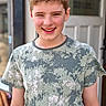 boy, child, smiling, winking, short_hair, camouflage_shirt, standing, casual, outdoor, door, wall, portrait, face, skin, person, happy, young, light, daylight, background_blur