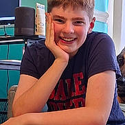 James joined the competition — help win amazing prizes! child, smiling, portrait, indoor, cat, home, furniture, desk, computer, speaker, window, colorful, short_hair, t_shirt, blue_shorts, seated, hand_on_cheek, happy, face, photography
