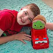 James is registered to the contest to win money with this photo: child, boy, smile, red_shirt, toy_truck, plush_toy, carpet, indoor, happy, face, hand, head, play, childhood, cute, car, toy, floor, casual, portrait