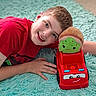 child, boy, smile, red_shirt, toy_truck, plush_toy, carpet, indoor, happy, face, hand, head, play, childhood, cute, car, toy, floor, casual, portrait