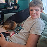 James is registered to the contest to win money with this photo: child, boy, smiling, portrait, indoor, couch, dogs, pet, gaming, game_controller, living_room, home, window, television, computer_setup, casual_clothing, t_shirt, seated, relaxed, family_pet