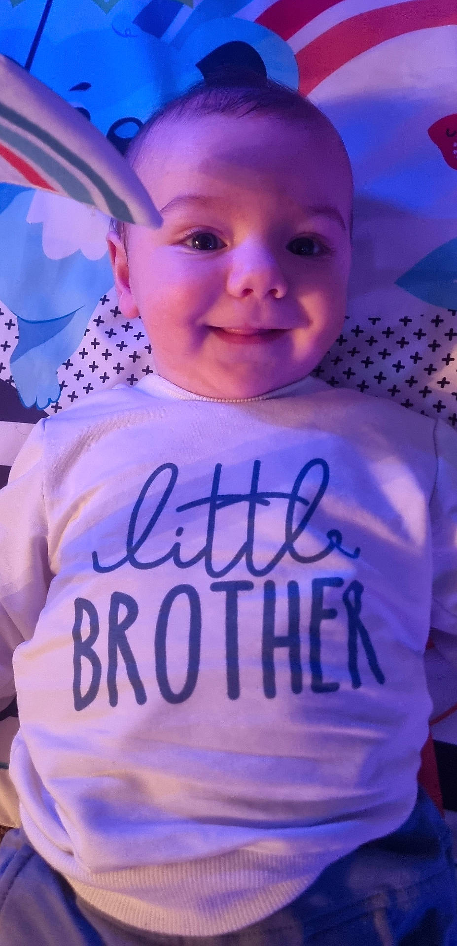 Orlando is registered to the contest to win money with this photo: baby, blue_light, child, closeup, clothing, colorful_background, cute, face, happy, indoors, infant, little_brother, lying_down, pattern, person, playmat, portrait, shirt, smiling, text