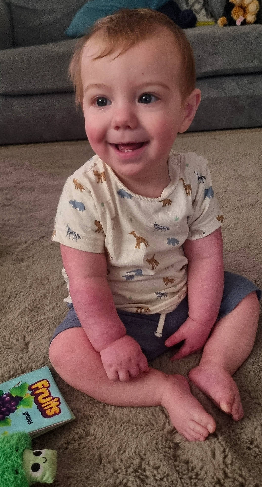 animal_print, carpet, child, cute, face, floor, furniture, happy, home, indoor, person, play, relaxed, shirt, shorts, sitting, smiling, toddler, toy, young_child