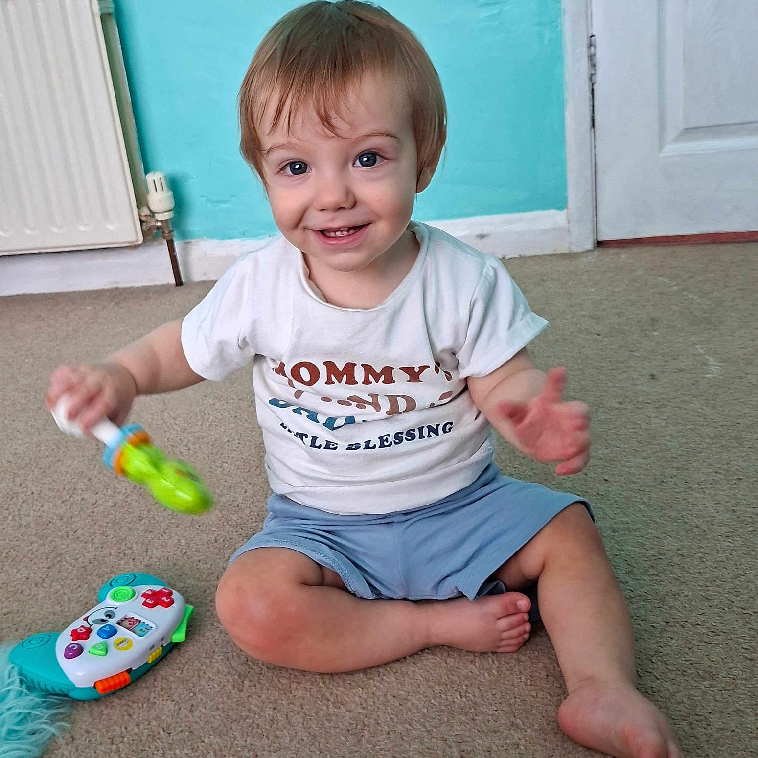 Orlando is registered to the contest to win money with this photo: toddler, child, toy, carpet, smile, sitting, indoor, white_tshirt, blue_shorts, radiator, door, wall, floor, happy, playtime, person, baby, cute, furniture, home